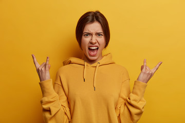 Irritated carefree mad young woman yells and feels excited, makes rock n roll gesture, goes wild while listens loud music, wears sweatshirt, isolated over yellow background. Rock this world!