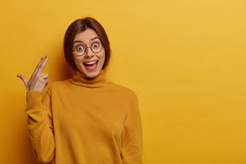 Funny teenage girl makes suicide gesture shoots in temple keeps mouth opened, has positive joyful gaze at camera, pretends commiting suicide, killing herself, wears round transparent glasses, poloneck - obrazy, fototapety, plakaty
