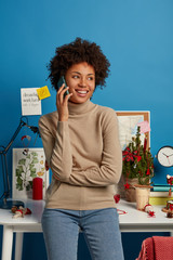 Photo of pleased Afro American freelancer stands in own cabinet at home, has phone conversation, looks aside with broad smile, poses near desktop with desk lamp, decorated fir tree, blue background