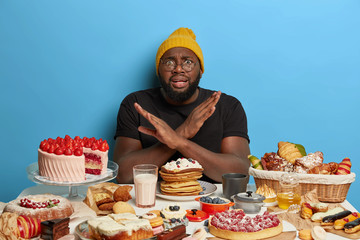African American man crosses arms over chest, makes denial gesture, refuses eat sugary products,...