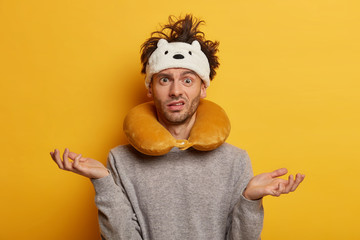 Confused man traveller has messy hairstyle, wears blindfold and travel pillow, spreads palms...