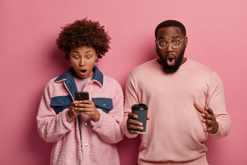 Stupefied emotional woman stares at smartphone display, gets strange text message, surprised bearded man drinks takeaway coffee, find out shocking news, stand against pink pastel background.