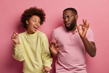 Overjoyed woman and man with dark skin have upbeat mood, dance at disco party, raise arms and move with rhythm of music, wear casual clothes, isolated on pink background. People, fun, joy concept