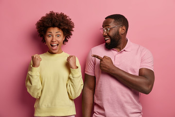 Happy black man with thick beard points at triumphing woman with clenched fists, celebrates...