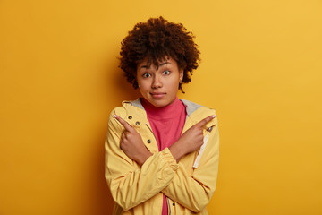 Confused dark skinned woman crosses arms over chest, chooses both ways left and right, makes choice, shows what decision better, wears anorak, isolated over yellow background. Two choices, options