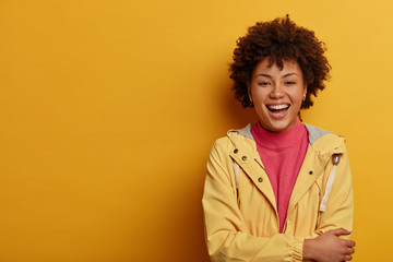 Obraz na płótnie Canvas Enthusiastic hilarious woman with Afro hairstyle, laughs out loud, imagines funny situation, keeps hands crossed over chest, dressed casually, stands against yellow wall, copy space, smiles happily