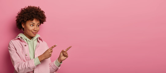 Friendly looking cheerful woman with Afro hairstyle indicates with index fingers aside, shows copy space for your advertising content, demonstrates nice promo, wears casual clothes, isolated on pink