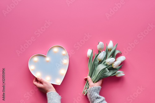 Flat lay, top view on pink, Mothers day or Valentine's holiday. female woman hands holding bunch of white tulips, Spring flowers. Lightboard in heart shape with illuminated lights or small lamps.
