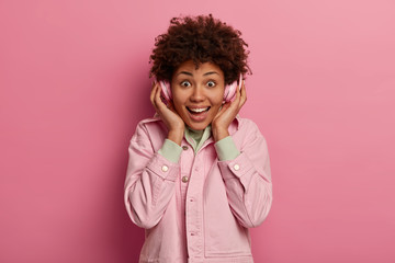 Positive teenager spends free time listening music, wears stylish pastel pink jacket, has toothy smile and giggles positively, being amused, isolated over rosy studio wall, hears popular hit on radio