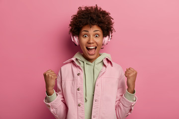 Excited overemotive lucky Afro American woman finds out she won, shows fist pump, exclaims joyfully, gets approval, listens loud favourite music in headphones, keeps mouth opened, celebrates triumph
