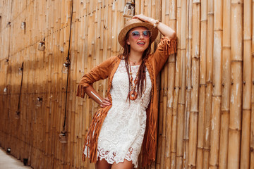 Lady dressed in boho style, straw hat, cape, bracelets and pendant made of eco wood, glasses, flash tattoos. Standing on the background of a bamboo wall, summer vacation fashion