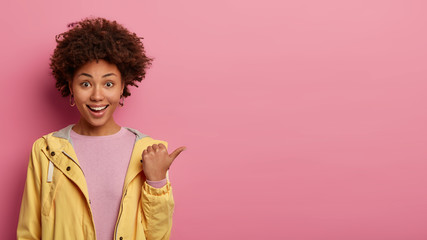 Positive curly haired woman points thumb aside, has glad expression, demonstrates something interesting, wears casual jacket, poses over rosy wall with empty space. People and advertisement.
