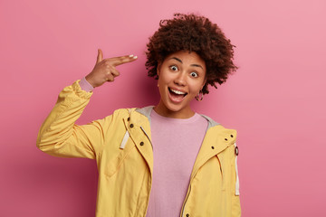 Shot of playful curly woman makes finger gun pistol gesture, shoots in temple, has joyful...