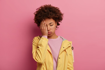 Fototapeta na wymiar Tired female teenager sighs from unhappiness, covers face with palm, closes eyes and suffers from headache, being overworked, has sleepy expression wears casual anorak, stand over rosy studio wall.