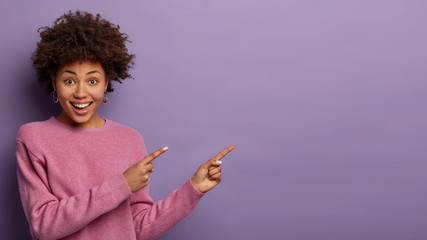 Joyful pretty girl with Afro haircut recommends you nice thing on sale, demonstrates blank space for advertisement, laughs sincerely, wears casual jumper, gestures over purple background. Look right
