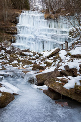 Beautiful, scenic, frozen Tupavica waterfall with hanging icicles and mountain creek with orange rocks and wooden bench and picnic table