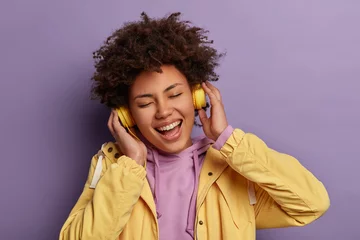 Gordijnen Close up portrait of glad funny curly haired woman listens favourite music with headphones, forgets about all troubles, enjoys loud tune, keeps eyes closed, smiles broadly, isolated on purple wall © Wayhome Studio