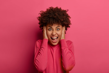 Fototapeta na wymiar Outraged crazy emotional woman screams with negative emotions, being in panic, yells and expresses hate, anger or rage, grabs head, wears crimson jumper, isolated on pink background, feels mad