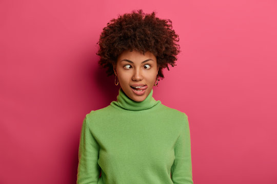 Curly haired female crosses eyes and sticks out tongue, feels like crazy clown, wears green turtleneck, makes grimace, foolishes around, stands against pink background, shows hilarious funny face