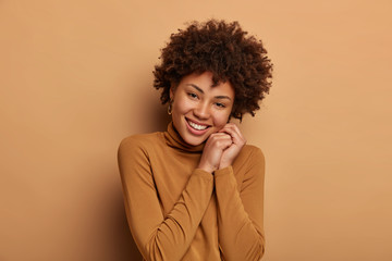 Fototapeta na wymiar People and pleasant emotions concept. Gentle positive woman tilts head, smiles with delight, gazes tenderly at camera, feels happiness, wears casual jumper, has cheerful grin, isolated on beige wall
