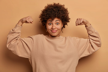 Strong powerful curly haired woman has cheerful playful expression, raises arms and shows muscles,...