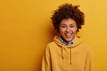Studio shot of joyful teenager laughs sincerely, wears casual hoodie, expresses positive emotions, has casual funny talk with interlocutor, stands over yellow wall, copy space left for your text