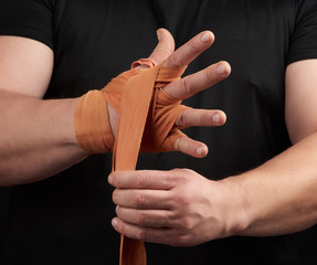 athlete stand in black clothes and wrap his hands in red textile elastic orange bandage