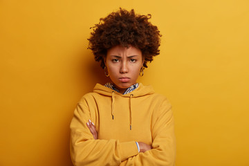 Fototapeta na wymiar Feeling offended and displeased. Dissatisfied Afro American woman looks from under forehead, crosses hands over chest, wears casual hoodie, isolated over yellow background. I am not trusting you