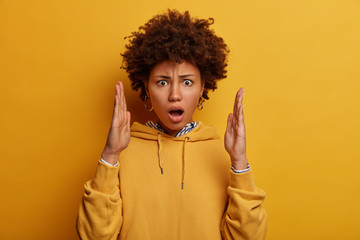 Isolated shot of impressed shocked woman makes big size gesture, shows something huge with dissatisfaction, keeps jaw dropped from surprisement, dressed in yellow sweatshirt, explains shape of item