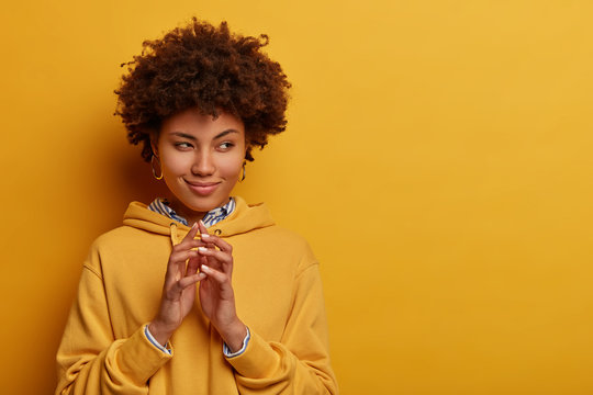 Mysterious dark skinned woman has interesting plan in mind, steeps fingers and looks aside, intention doing something, everything goes according scheme, has curious cunning expression, wears hoodie