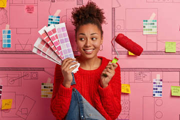 Cheerful woman holds palette and red paint roller, looks aside, has curly combed hair, poses...