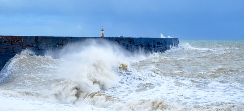 A harbour wall with a rough sea crashing against it and a lighthouse in the background, Newhaven harbour, Sussex, England
