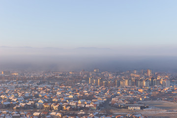 Wide view of misty cityscape with buildings and houses lighten by golden sunrise sun and rooftops covered by first snow