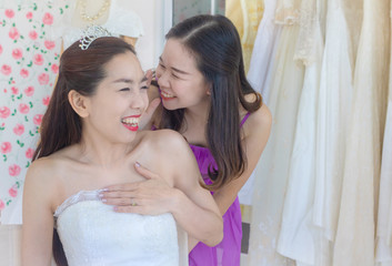 Young Asian bride and bridesmaid are whispering about something and laughing with wedding dress background.They are cheerful and enjoy in wedding day.happy,excited,Photo concept marry and ceremony.