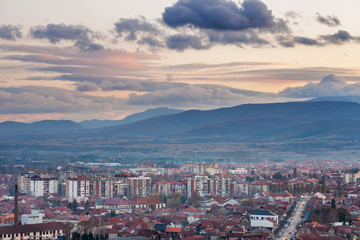 Pirot, Serbia cityscape viewed from a vantage point during a sunset with foreground buildings and city lights and distant mountain layers under colorful sky 