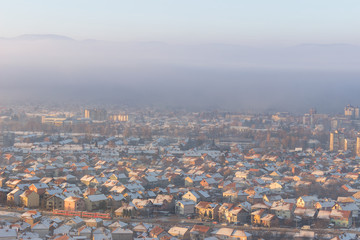 Obraz premium Red train passing in front of misty cityscape with buildings lighten by golden sunrise and rooftops covered by first snow