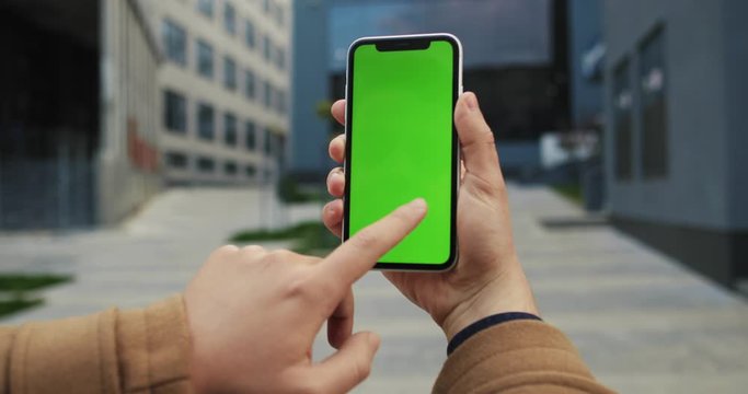 Caucasian male hand holding black smartphone vertically with green screen like scrolling pictures on it on the street. Close up. Outside. Chroma key.