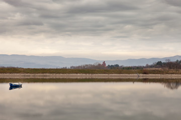Unique view across calm, reflective Kragujevac lake with old, blue boat during a cloudy, moody day, on memorial museum 21st October on Sumarice in the distance