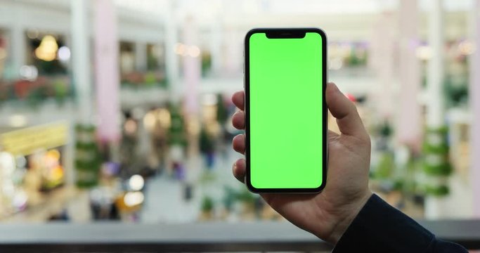 Close up of the black vertical smartphone with chroma key on its screen in hand of Caucasian man while he scrolling pictures or pages on it at the shopping mall.