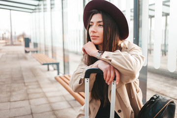 Pensive stylish girl in a coat and hat sits at a stop waiting for public transport