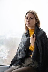 lovely young girl in an orange sweater under a plaid sits resting on a windowsill by a large window