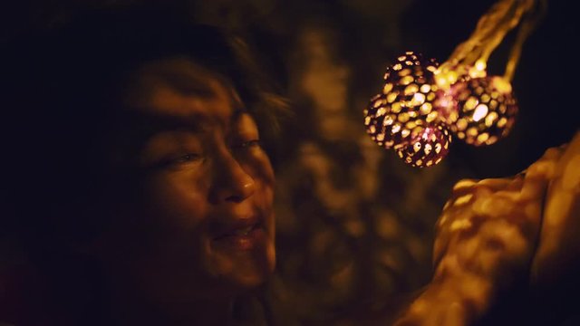Magic hot light from lanterns falls on the woman eyes. The naked adult sexy caucasian girl lies at night on the bed and holds warm lamps in her hands. Christmas toy in black background. Indoor.