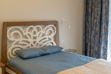 The interior in blue-beige of the bedroom. Part of a big original bed with wood carvings. Next to it is a lamp and electrical plugs on the wall,  a glass table and a window with curtains