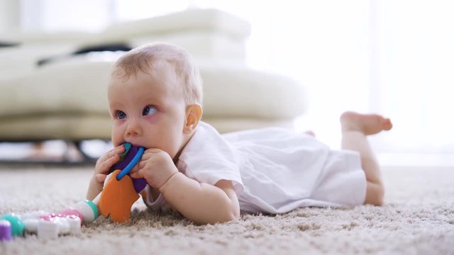 Cute baby girl playing and biting her toy while lying on the fur carpet in living room at home. Shot in 4k resolution