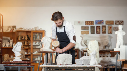 Bearded craftsman works in white stone carving with a chisel. Creative workshop with works of art. - obrazy, fototapety, plakaty