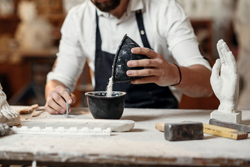 Unrecognizable professional stonemason with well-maintained hands making glue in special vessel for his handmade sculpture