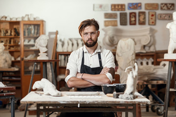 Portrait of attractive joyful bearded male sculptor which sitting on the workshop interior background and looks at the camera with smile. - obrazy, fototapety, plakaty