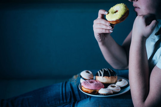 Overweight Depressed Woman Laying On Sofa Eating Sugary Food Watching TV. Sugar Addiction, Unhealthy Lifestyle, Weight Gain, Dietary, Healthcare And Medical Concept.