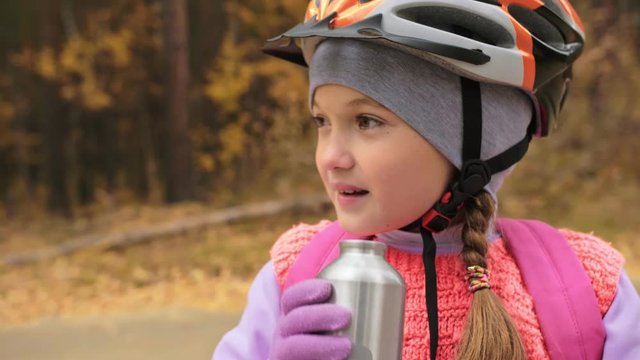 Kid drink water from an aluminum flask bottle. One caucasian children rides bike road in autumn park. Little girl riding black orange mtb cycle in forest. Biker motion ride with backpack and helmet.