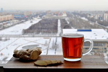 A transparent glass mug of hot black tea and sliced ​​ginger root close-up stand on a wooden surface against a blurry background of the winter panorama of the city of Togliatti.